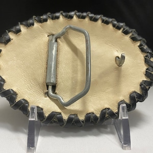 Peut inclure: Une boucle de ceinture en cuir blanc avec une bordure en cuir noir. La boucle est de forme ovale et poss&egrave;de un fermoir en m&eacute;tal.