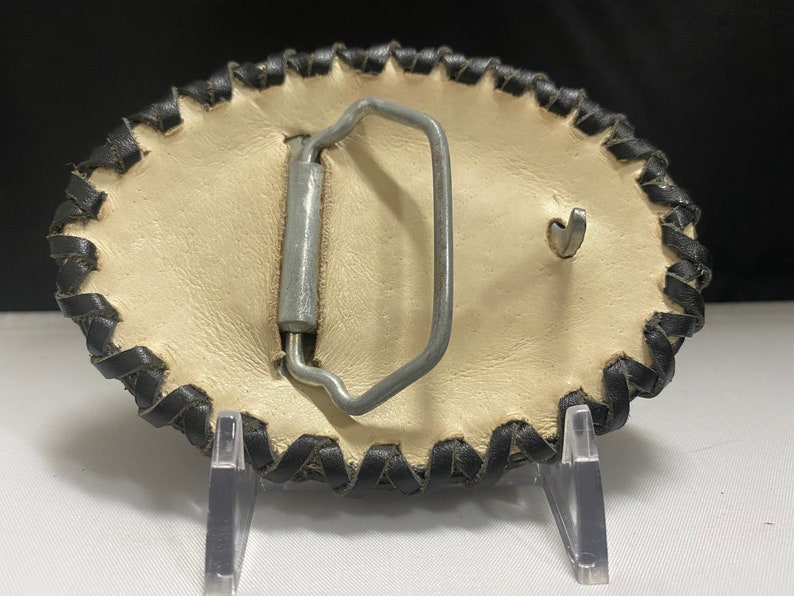 Peut inclure: Une boucle de ceinture en cuir blanc avec une bordure en cuir noir. La boucle est de forme ovale et poss&egrave;de un fermoir en m&eacute;tal.