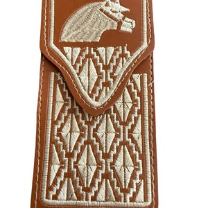 May include: A brown leather wallet with white embroidered horse head and geometric pattern. The wallet has a flap closure and white stitching around the edges. The design is reminiscent of Western or equestrian themes.