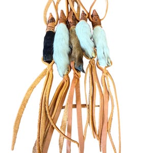 May include: A collection of six leather and fur charms. Each charm features a small animal paw with white, brown, and black fur, attached to a leather strap. The charms are suspended from a loop of leather.