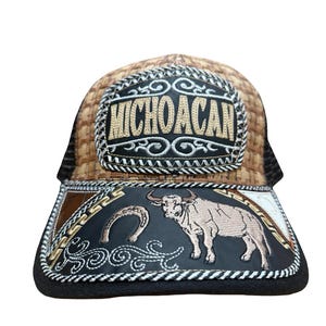 Peut inclure: Casquette de camionneur avec un dessus marron tissé et un dos en filet noir. L'avant présente un rectangle noir avec le mot "MICHOACAN" brodé en or. En dessous, un motif de taureau et de fer à cheval.