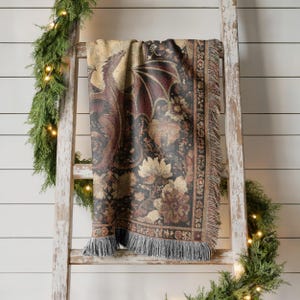 May include: A tapestry throw blanket draped over a wooden ladder. The blanket features a dragon design with floral accents in shades of brown, black, and gold. The blanket has a fringed edge.
