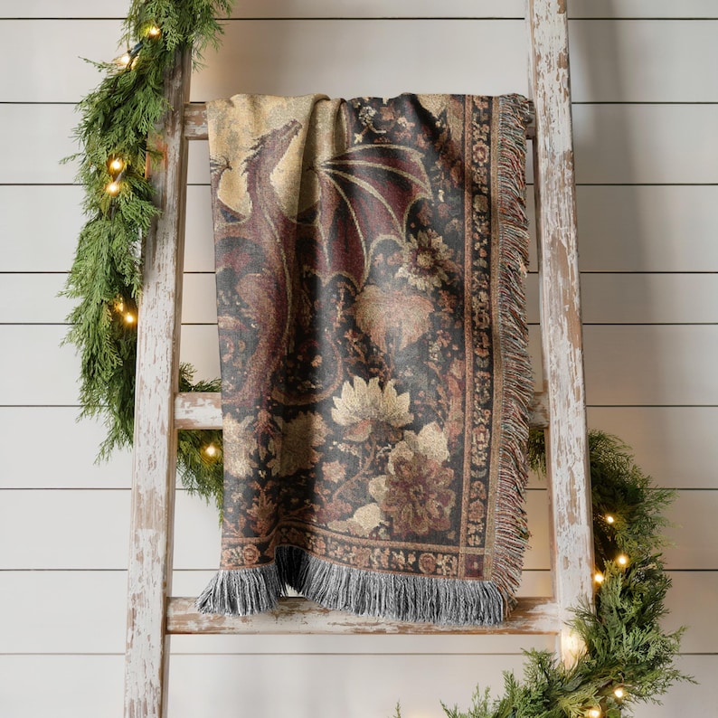 May include: A tapestry throw blanket draped over a wooden ladder. The blanket features a dragon design with floral accents in shades of brown, black, and gold. The blanket has a fringed edge.