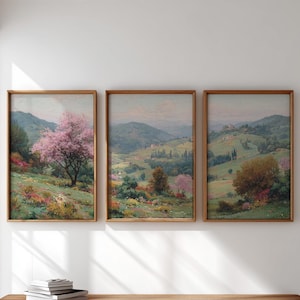 May include: Three framed landscape paintings. Each features a pastoral scene with rolling green hills, trees, and colorful flowers. The frames are a warm brown color, and the artwork evokes a sense of tranquility.