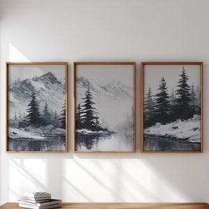 May include: Three framed landscape art prints. Each print features a monochrome mountain scene with evergreen trees and a reflective body of water. The frames are made of light brown wood.