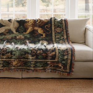 May include: A decorative throw blanket featuring a detailed design of mythical creatures, including a griffin and a lion, in shades of gold, brown, and green on a dark background. The blanket has a fringed edge and is draped over a beige sofa.