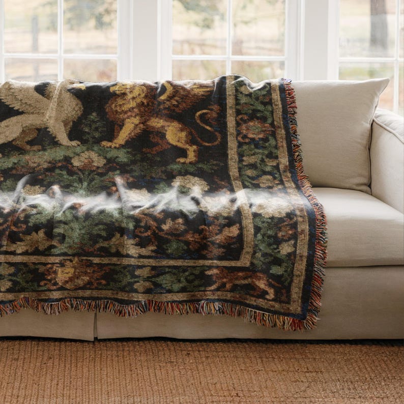 May include: A decorative throw blanket featuring a detailed design of mythical creatures, including a griffin and a lion, in shades of gold, brown, and green on a dark background. The blanket has a fringed edge and is draped over a beige sofa.