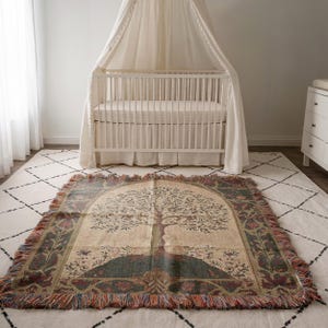 Peut inclure: Une couverture tiss&eacute;e avec un motif d'arbre, fond beige, colline verte et bordure d&eacute;corative. Le plaid est pos&eacute; sur un tapis blanc devant un lit b&eacute;b&eacute; blanc.