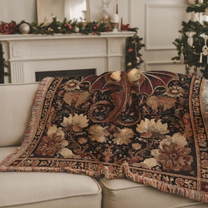 May include: A decorative throw blanket featuring a dragon design with a black background and floral accents. The blanket has a fringed edge and is draped over a beige sofa. The dragon is in shades of red and gold.
