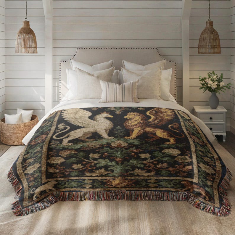 May include: A decorative throw blanket featuring a griffin and a lion on a dark background with floral accents. The blanket has a fringed edge with red, brown, and blue threads. The blanket is displayed on a bed with white pillows.