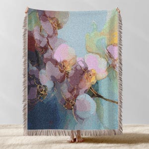 May include: A throw blanket featuring a floral design with pink, purple, and gold accents on a blue background. The artwork depicts orchid-like flowers with gold detailing. The blanket has a fringed edge.