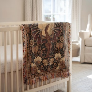 May include: A tapestry throw blanket with a dragon design draped over a white crib. The blanket features a dragon with outstretched wings in shades of brown and red, set against a dark floral background. The edges have a fringed trim.