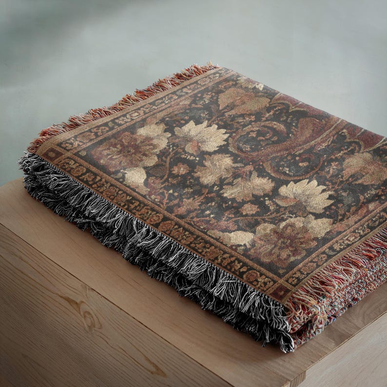 May include: A folded tapestry throw blanket with a floral and paisley pattern in shades of brown, gold, and burgundy. The blanket has a fringed edge in black and red, and is resting on a wooden surface.