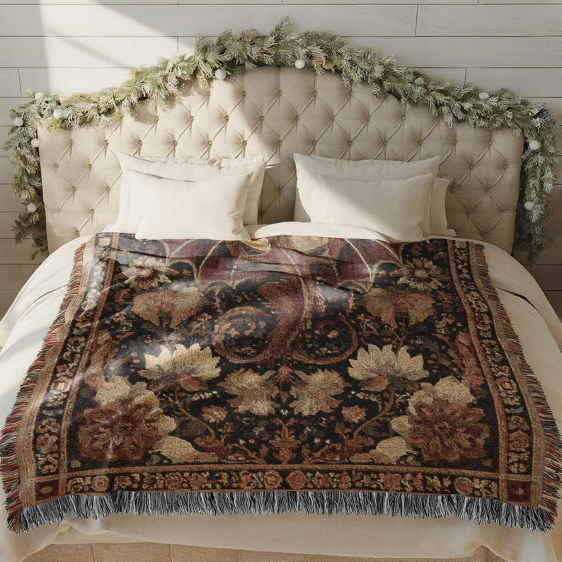 May include: A decorative throw blanket with a floral pattern in shades of black, brown, and gold, fringed edges, and a detailed border. The blanket is displayed on a bed with white linens and a tufted headboard.