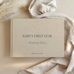 May include: A beige linen-covered baby's first year memory book. The book's cover features the text "BABY'S FIRST YEAR", the name "Penelope Rose", and the date "June 24, 2025" in a dark gray font. The book is set against a neutral background with fabric and dried plants.