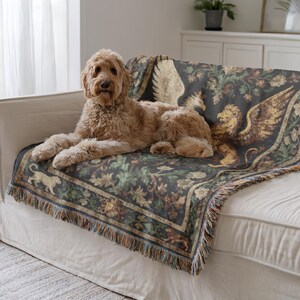 May include: A golden doodle dog rests on a white sofa, draped with a decorative tapestry throw. The throw features a dark background with a repeating pattern of griffins, foliage, and a border with fringe.