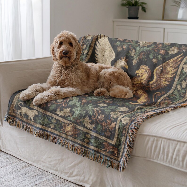 May include: A golden doodle dog rests on a white sofa, draped with a decorative tapestry throw. The throw features a dark background with a repeating pattern of griffins, foliage, and a border with fringe.