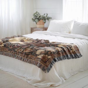 May include: A decorative throw blanket with a floral design in shades of brown, gold, and burgundy, fringed edges, draped over a white bed. The blanket features a detailed pattern and adds a touch of elegance to the bedroom decor.