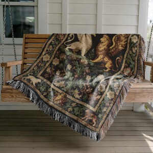 May include: A woven tapestry throw blanket featuring a griffin and lion design in shades of gold, tan, and brown on a black background. The blanket has a fringed edge and is draped over a wooden swing.