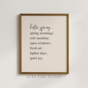 May include: A framed print with a gold-colored frame. The print has a beige background with black text that reads "hello spring... spring mornings. soft sunshine. open windows. fresh air. lighter days. quiet joy." The bottom of the print reads "LUXE VIBE STUDIO."