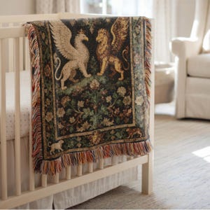 May include: A woven tapestry blanket draped over a white crib. The blanket features a design of a griffin and a winged lion, with floral and foliage details in shades of green, brown, and gold. The blanket has a fringed edge.