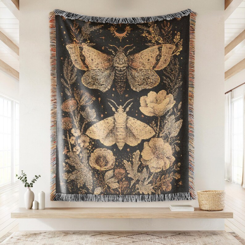 May include: Black throw blanket featuring a moth and floral design in gold and tan. The blanket has a fringed edge and is displayed on a wall. The design includes two moths, flowers, and foliage.