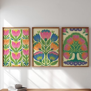 Może przedstawiać: Trzy oprawione grafiki kwiatowe. Grafiki przedstawiają kolorowe, stylizowane kwiaty w kolorze różowym, pomarańczowym i niebieskim na kremowym tle. Ramy są drewniane. Grafika to dekoracja ścienna.
