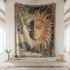 May include: Woven tapestry featuring a celestial design with the sun and moon. The artwork depicts the sun in shades of orange and yellow, and the moon in cream and gray, set against a dark starry background. The tapestry has a fringed edge.