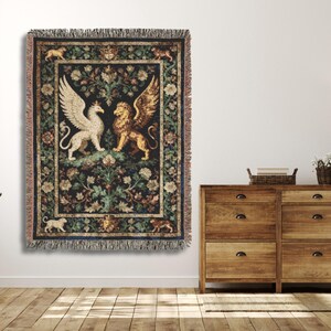 May include: A tapestry throw blanket featuring a central design of a white griffin and a golden lion with wings, set against a dark background with floral and foliage patterns. The blanket has a fringed edge and is hung on a white wall.