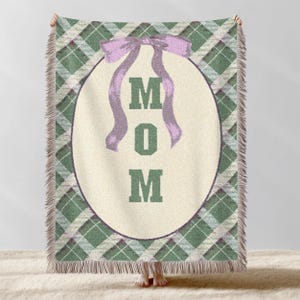 May include: A green and white plaid throw blanket with a cream oval in the centre. The oval features the word "MOM" in green block letters, with a lavender bow at the top. The blanket has a fringed edge.
