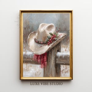 May include: A framed painting depicts a cowboy hat adorned with holly and berries, resting on a snow-covered fence. A red scarf hangs below the hat. The artwork is set against a snowy, wintery backdrop, with the text "LUXE VIBE STUDIO" at the bottom.