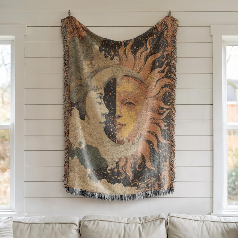 May include: A woven tapestry featuring a celestial design of the sun and moon. The artwork is in shades of beige, gold, and black, with a fringed edge. The tapestry is hanging on a white wall.