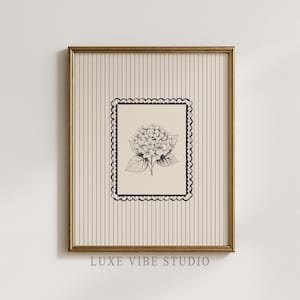 Op de afbeelding: Een ingelijste bloemenkunstprint met een zwart-wit illustratie van een hortensia. Het kunstwerk staat tegen een crèmekleurige achtergrond met verticale strepen en een decoratieve zwarte rand. De lijst is goudkleurig en de tekst "LUXE VIBE STUDIO" staat onderaan.