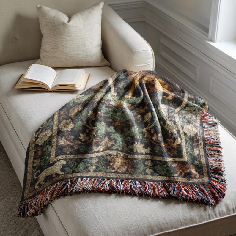 May include: A decorative throw blanket with a forest-themed pattern, featuring shades of green, brown, and gold. The blanket has a fringed edge with multiple colors and is draped over a chaise lounge. An open book and a pillow are also visible.