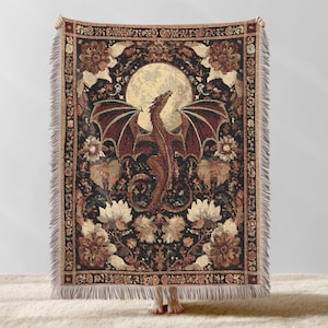 May include: A tapestry throw blanket featuring a dragon with outstretched wings against a full moon. The dragon is brown and red, surrounded by floral patterns in shades of gold, brown, and cream, set against a black background. The blanket has a fringed edge.