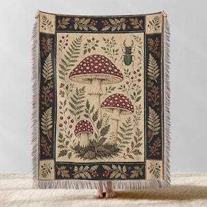 May include: A woven throw blanket featuring a central cream-colored panel with three red-capped mushrooms, green ferns, and a beetle. The design is framed by a dark border with botanical motifs and finished with fringed edges on all sides.