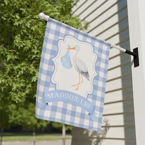 May include: A blue and white gingham flag with a stork carrying a baby in a blue bundle. The flag has the name "MADDOX LEE" in a banner across the bottom. The flag is hanging outside on a white wall.