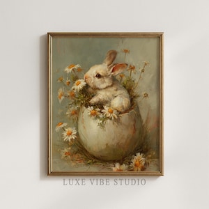 Puede incluir: Una impresión enmarcada de estilo vintage que presenta un conejito blanco en un cascarón de huevo agrietado, rodeado de margaritas. La obra de arte tiene una paleta de colores pastel y un marco dorado. El texto "LUXE VIBE STUDIO" está impreso en la parte inferior.