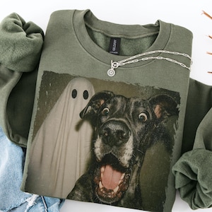 May include: An olive green sweatshirt featuring a graphic of a surprised dog and a ghost. A silver necklace with a circular pendant is draped across the sweatshirt. The sweatshirt is folded, with a portion of a blue denim garment visible.