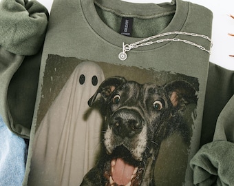 Può includere: Una felpa verde oliva con una grafica di un cane sorpreso e un fantasma. Una collana d'argento con un ciondolo circolare è drappeggiata sulla felpa. La felpa è piegata, con una porzione di un capo in denim blu visibile.