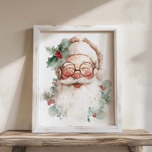 May include: Watercolor print of Santa Claus in a white distressed frame. Santa is depicted with a long white beard, round glasses, and a red nose. He is surrounded by holly and eucalyptus. The artwork is displayed on a wooden surface.