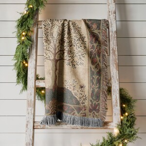 Peut inclure: Une couverture tiss&eacute;e avec un motif d'arbre dans les tons beige, marron et vert est drap&eacute;e sur une &eacute;chelle en bois rustique. La couverture a un bord &agrave; franges et une bordure d&eacute;corative. L'arri&egrave;re-plan est un mur blanc &agrave; planches horizontales.