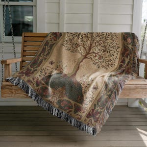 Peut inclure: Une couverture tiss&eacute;e avec un motif d'arbre drap&eacute;e sur une balan&ccedil;oire de porche en bois. La couverture pr&eacute;sente un fond beige avec une illustration d&eacute;taill&eacute;e d'arbre en marron et vert, encadr&eacute;e par une bordure d&eacute;corative avec des franges.