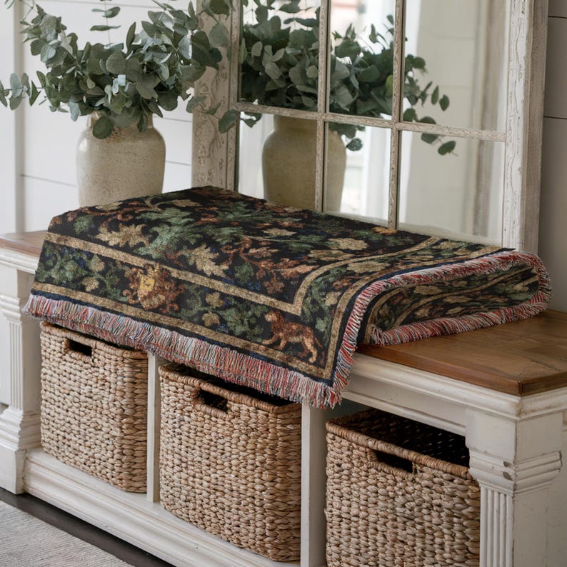 May include: A folded tapestry throw blanket with a dark background and a detailed pattern of foliage and animals. The blanket has a fringed edge with red, blue, and tan threads. The throw is placed on a wooden bench with three woven baskets below.