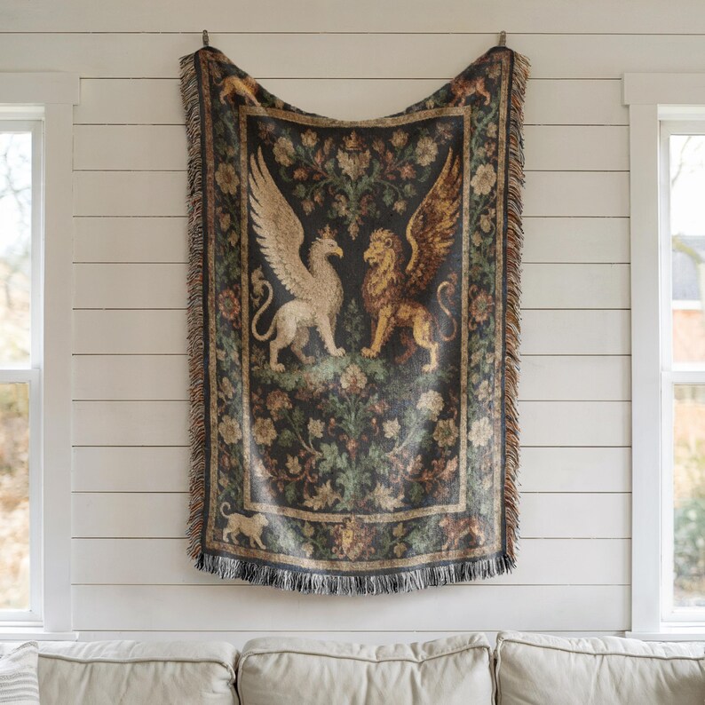 May include: A tapestry-style throw blanket featuring a central design of a winged griffin and a winged lion facing each other. The blanket has a dark background with floral and foliage patterns, and a fringed edge. The color palette includes shades of black, beige, and gold.