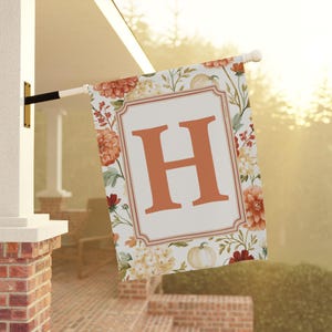 May include: A rectangular garden flag featuring a large orange letter H centered within a decorative frame. The background is decorated with a pattern of orange and yellow flowers, green foliage, and small white pumpkins on a white fabric surface.