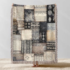 May include: A patchwork throw blanket with a variety of patterned squares in shades of beige, black, and gray. The blanket has a fringed edge and a mix of floral, geometric, and paisley designs. The blanket is held up by a person.
