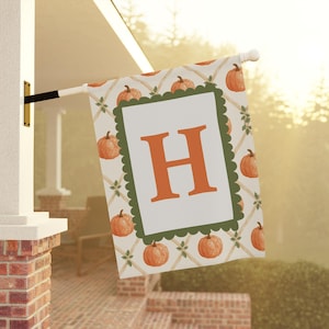May include: A rectangular garden flag featuring a white background with a repeating pattern of orange pumpkins and tan lattice lines. A central white rectangle with a scalloped dark green border displays a large, bold, orange letter H.