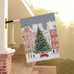May include: A decorative Christmas flag featuring a watercolor painting of a snowy village scene. The design includes a decorated Christmas tree, red buildings, a red car, and people walking in the snow. The flag is mounted on a white pole.