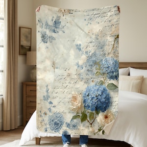 May include: A large, rectangular throw blanket featuring a vintage floral design. The blanket showcases blue hydrangeas and cream-colored roses against a backdrop of faded script and watercolor washes. The color palette includes shades of blue, cream, and beige.